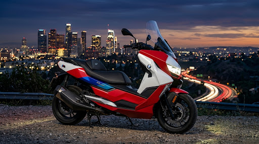 The red, white and blue BMW C 400 GT parked at a hillside overlook at dusk, with a city skyline and light trails behind it.